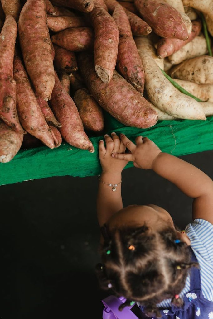 A young child curiously reaches for fresh sweet potatoes at an outdoor market stall.