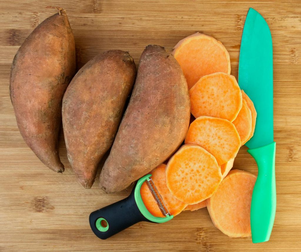 Close-up of sweet potatoes, sliced with a green knife and peeler on a wooden surface.